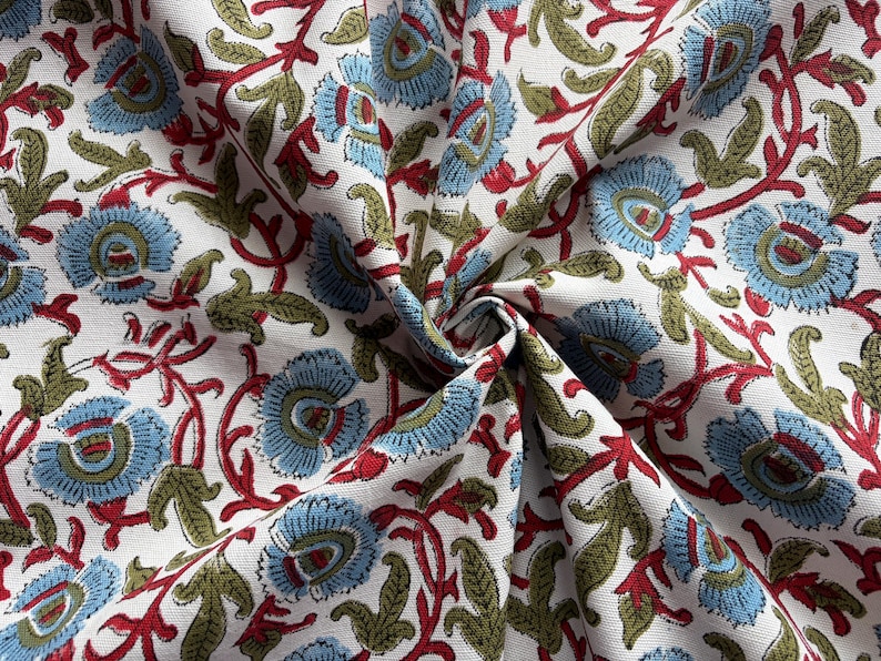 May include: A white fabric featuring a repeating floral pattern with light blue petals, olive green leaves, and red stems. The textile is gathered in the center, creating folds that highlight the intricate botanical print across the surface.