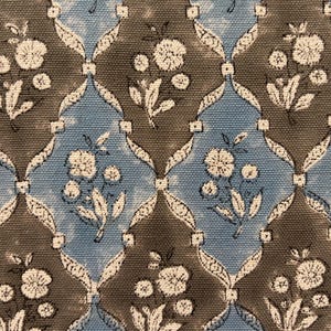 Op de afbeelding: Close-up van een stof met een herhalend bloemenpatroon. Het ontwerp toont witte bloemen en bladeren op een blauwe en grijze achtergrond. Het patroon is gerangschikt in een roosterachtige structuur, waardoor een visueel aantrekkelijk textiel ontstaat.