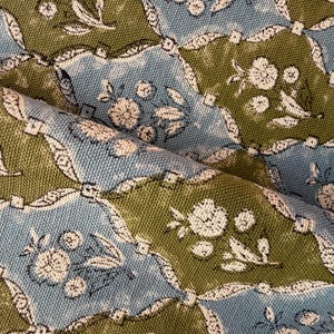 May include: Close-up of a patterned fabric with a repeating design. The pattern features floral and leaf motifs in white against a backdrop of blue and olive green. The fabric is folded, showing texture and depth.