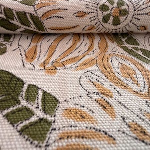 May include: Close-up of a cream-colored fabric with a repeating floral pattern. The design features olive green leaves and tan-colored floral elements, outlined in black. The fabric has a textured appearance.