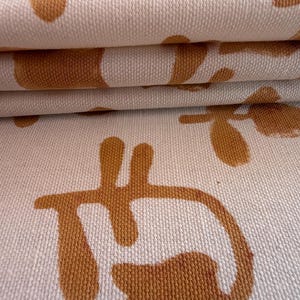 May include: Close-up of folded fabric with a cream-colored background and a repeating pattern of brown abstract shapes. The fabric has a textured appearance, with visible weave details. The pattern includes floral and geometric elements.