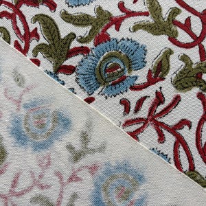 May include: A white fabric featuring a repeating floral pattern with light blue petals, olive green leaves, and dark red stems. The textile shows a clear, vibrant print on the upper right section and a faded, muted version on the lower left.