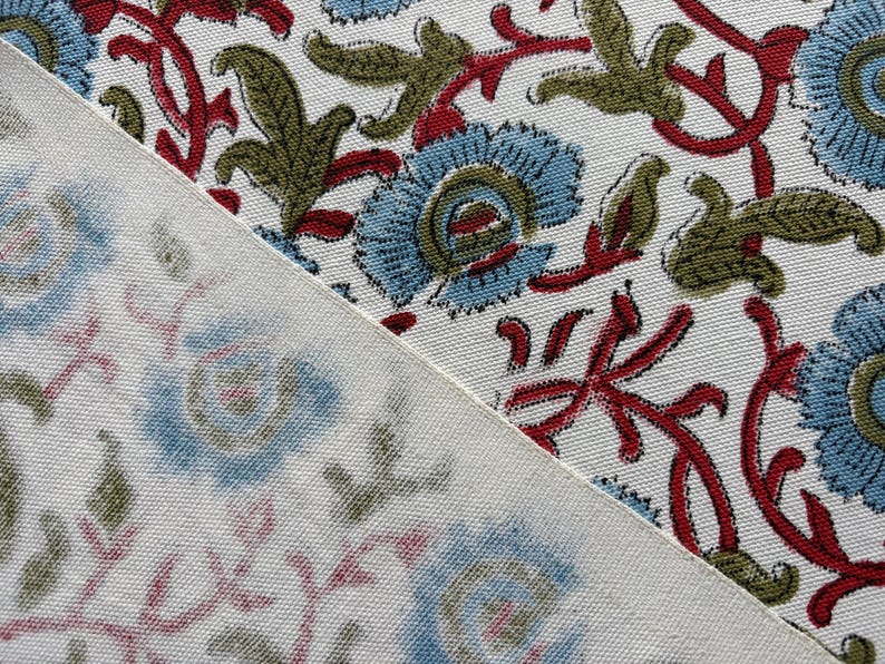 May include: A white fabric featuring a repeating floral pattern with light blue petals, olive green leaves, and dark red stems. The textile shows a clear, vibrant print on the upper right section and a faded, muted version on the lower left.