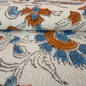Puede incluir: Primer plano de una tela doblada con un estampado floral repetitivo. El diseño presenta flores naranjas y azules con contornos negros sobre un fondo crema. La tela tiene una apariencia texturizada.