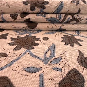 以下が含まれることがあります： 折りたたまれた生地のクローズアップ。花柄のデザインで、クリーム色の背景に茶色と青の花と葉が描かれています。生地は質感のある外観をしています。