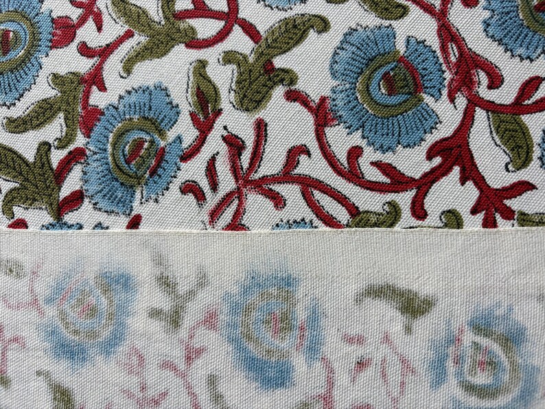 May include: A textile fabric featuring a repeating floral pattern with light blue petals, olive green leaves, and dark red stems on a white background. The lower section shows a faded, muted version of the same floral print on a cream-colored fabric.
