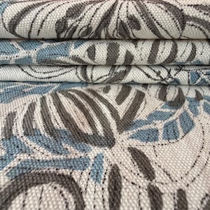 Puede incluir: Primer plano de tela doblada con un estampado floral. El diseño presenta hojas y flores estilizadas en tonos grises y azules sobre un fondo blanco. La tela parece ser un material tejido, posiblemente lino o algodón.