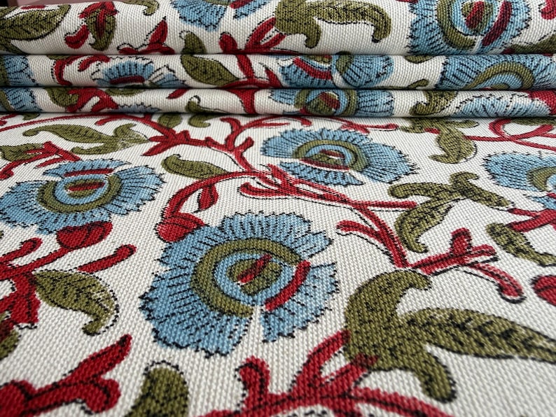 May include: A woven fabric featuring a repeating floral pattern on an off-white background. The design consists of light blue flowers with olive green centers, connected by vibrant red stems and olive green leaves, creating a textured surface.