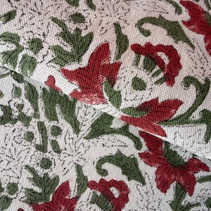 May include: Close-up of folded fabric with a repeating floral pattern. The design features red flowers and green leaves on a cream background. The fabric has a textured appearance.