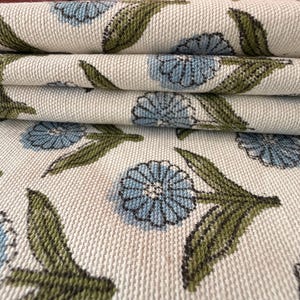 May include: Close-up of folded fabric with a repeating floral pattern. The design features light blue flowers with green leaves and stems on a cream-colored background. The fabric appears to be a woven material.