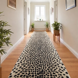 Può includere: Un lungo tappeto rettangolare con sfondo color crema e motivo a macchie nere, simile alla pelliccia di un leopardo, è steso in un corridoio con pavimenti in legno. Il corridoio ha pareti bianche, porte e opere d'arte incorniciate.