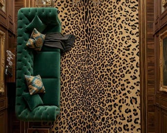 May include: A rectangular area rug featuring a tan and black leopard print pattern. The rug is positioned next to a deep green tufted velvet sofa adorned with patterned throw pillows and a dark gray throw blanket, set beneath a gold chandelier.