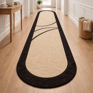 Könnte beinhalten: Ein ovaler Läufer in Beige und Schwarz. Der Teppich hat eine beige Mitte, einen schwarzen Rand und schwarze geschwungene Linien. Er liegt auf einem Holzboden in einem Flur. Die Abmessungen des Teppichs sind in Zentimetern.
