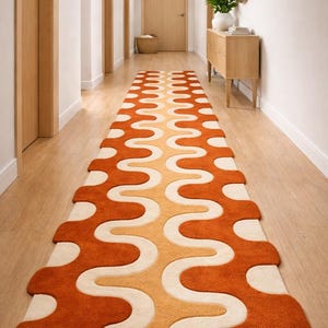 Op de afbeelding: Een lange, oranje, crème en gele patroonloper in een gang. De loper heeft een golvend ontwerp en is gecentreerd in de gang. De gang heeft licht houten vloeren en witte muren.