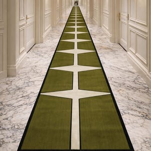 May include: A long hallway with white walls, doors, and marble flooring. A green and white patterned runner rug runs down the centre. The rug has a black border and a repeating star-like design. Wall sconces provide soft lighting.