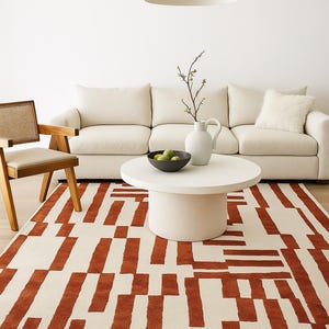 May include: A living room scene featuring a cream-colored sofa, a round white coffee table, and a wooden chair. A rug with a cream background and rust-colored geometric pattern covers the floor. A white pendant light hangs above the table.
