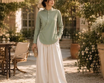 Puede incluir: Una túnica de manga larga en color verde salvia con cuello mao y cierre de botones laterales, combinada con pantalones de color crema de pernera ancha. El conjunto se complementa con un sombrero de paja de ala ancha con adornos florales.