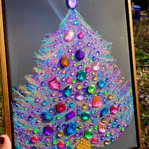 May include: A framed artwork on a black background featuring a stylized Christmas tree shape. The tree is decorated with a dense layer of purple and blue glitter, accented by numerous multi-colored faceted plastic gems and a gold sequined trunk.