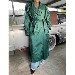 May include: A long, emerald green trench coat with a matching belt and double-breasted closure. The coat is worn over a brown sweater and blue jeans. The model is wearing dark red shoes. The coat appears to be made of a lightweight, water-resistant material.
