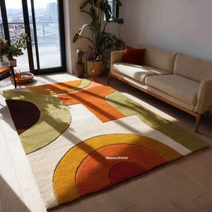 May include: A rectangular area rug with a geometric design in shades of orange, yellow, green, and brown. The rug features a cream-colored background with curved and rectangular shapes. The rug is placed on a wooden floor in a living room setting.