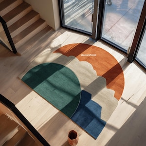 May include: A colorful, semi-circular rug with a mountain-like design. The rug features sections of green, beige, blue, and orange. The rug is on a light-colored wooden floor. The text "WeaversPointt" is visible on the rug.