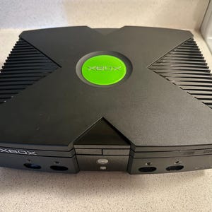 May include: Black Xbox console with a green circular power button. The console has a distinctive X-shaped design on top and the word "XBOX" is printed on the front. The console is sitting on a light-colored surface.
