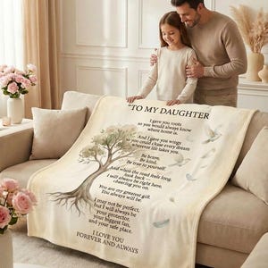 Op de afbeelding: Een beige deken met de tekst "TO MY DAUGHTER" en een boomillustratie. De deken heeft een zachte textuur en is over een bank gedrapeerd. De tekst bevat een liefdevolle boodschap. Een vader en dochter staan achter de deken.