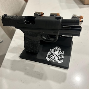 May include: A black handgun with a textured grip is displayed on a black stand. The stand features the Springfield Armory logo and the text "SINCE 1794". Two magazines with bullets are placed on top of the handgun.