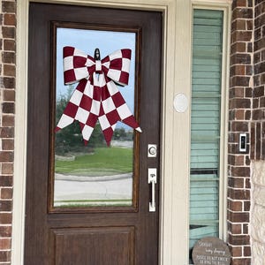 May include: A large decorative bow with a maroon and white checkered pattern hanging on a dark brown wooden door. Below the door window, a round wooden sign reads: SHHHH! baby sleeping PLEASE DO NOT KNOCK OR RING THE BELL thank you!