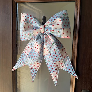 May include: A large, quilted fabric bow featuring a patchwork design of light blue gingham, white fabric with red strawberry prints, and light blue fabric with red lobster motifs. The bow has two main loops and two long, pointed tails hanging down.