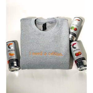 May include: A heather gray sweatshirt with the embroidered phrase "i need a celsius." The sweatshirt is surrounded by four cans of Celsius sparkling energy drink in various fruit flavors, including orange, kiwi, and strawberry.