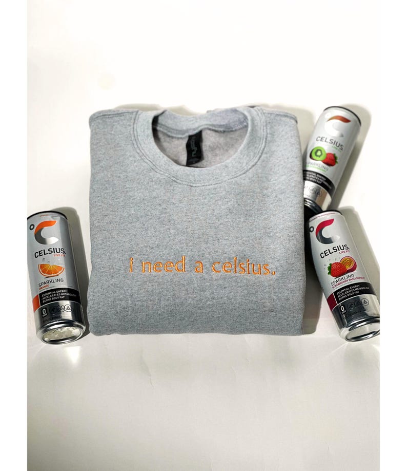 May include: A heather gray sweatshirt with the embroidered phrase "i need a celsius." The sweatshirt is surrounded by four cans of Celsius sparkling energy drink in various fruit flavors, including orange, kiwi, and strawberry.