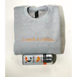 May include: A gray sweatshirt with the embroidered text "I need a celsius." A can of Celsius sparkling orange energy drink is below the sweatshirt. The can is silver with orange and black text and graphics.