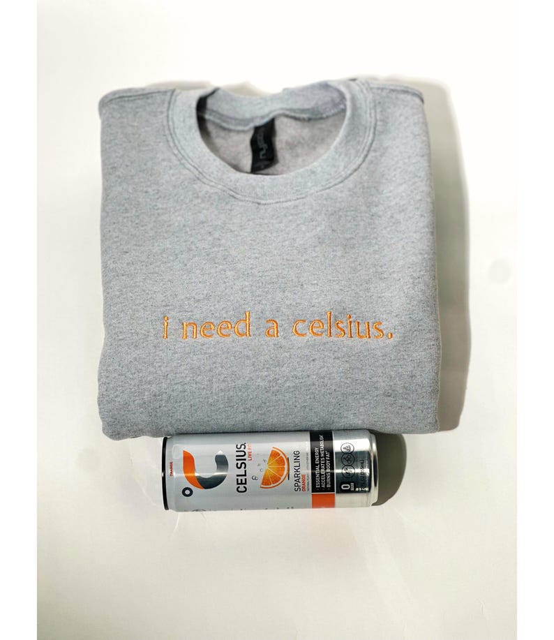 May include: A gray sweatshirt with the embroidered text "I need a celsius." A can of Celsius sparkling orange energy drink is below the sweatshirt. The can is silver with orange and black text and graphics.