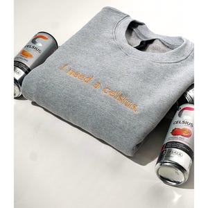 May include: A gray crewneck sweatshirt with the embroidered phrase "I need a celsius" in orange thread. The sweatshirt is surrounded by three cans of Celsius sparkling energy drink.