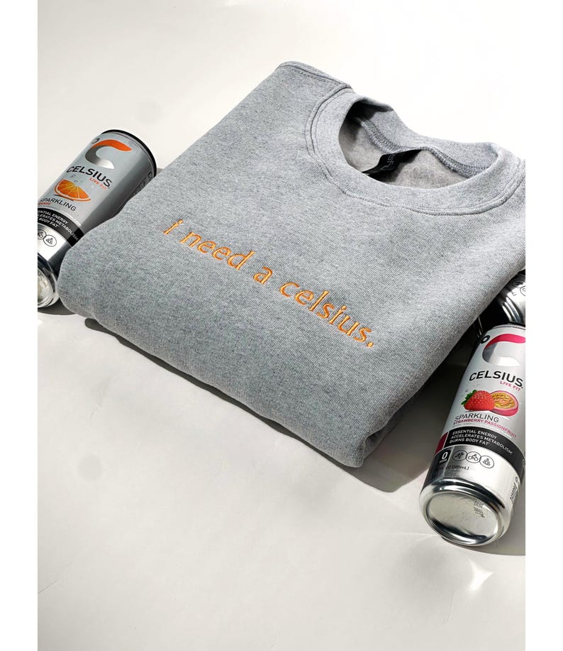 May include: A gray crewneck sweatshirt with the embroidered phrase "I need a celsius" in orange thread. The sweatshirt is surrounded by three cans of Celsius sparkling energy drink.