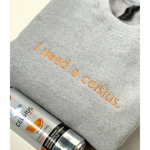 May include: A gray sweatshirt with the embroidered phrase "I need a celsius." is paired with a can of Celsius sparkling orange energy drink. The can is silver with orange accents and product information.