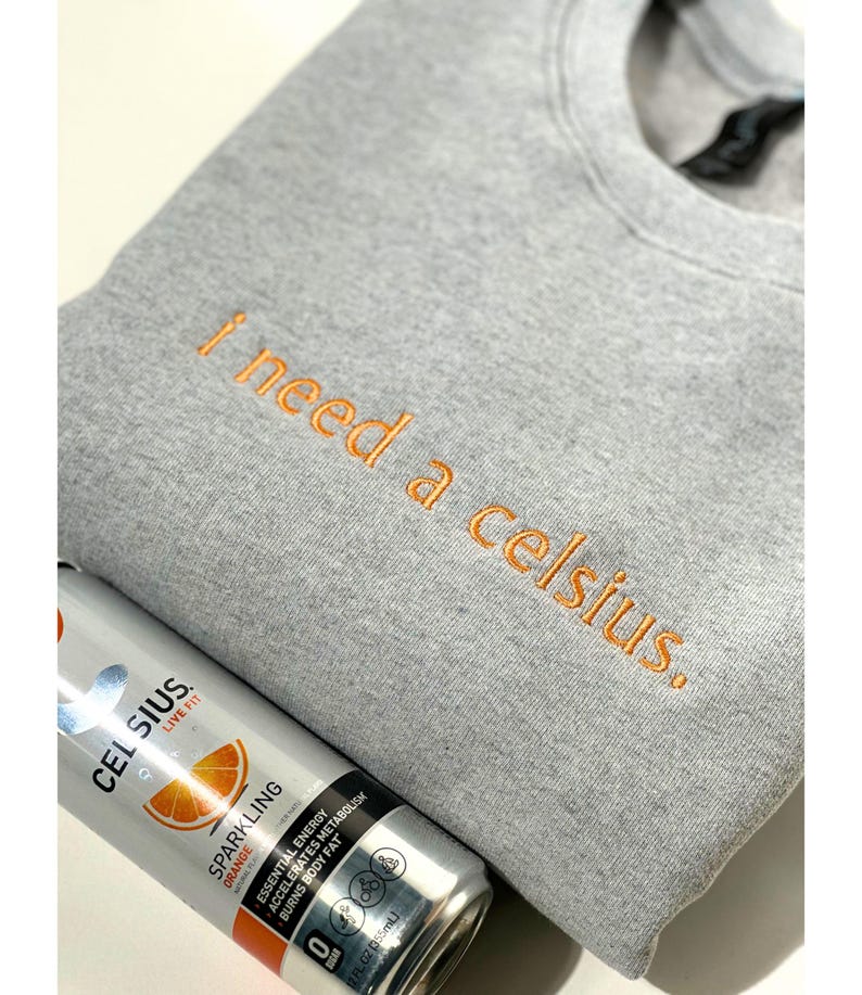 May include: A gray sweatshirt with the embroidered phrase "I need a celsius." is paired with a can of Celsius sparkling orange energy drink. The can is silver with orange accents and product information.