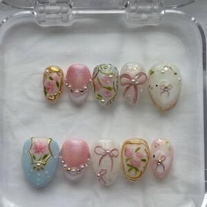 May include: A set of ten artificial nails in a clear plastic case. The nails feature various designs, including pink glitter, bows, floral patterns, and pearl accents. The color palette includes pink, gold, white, and light blue.