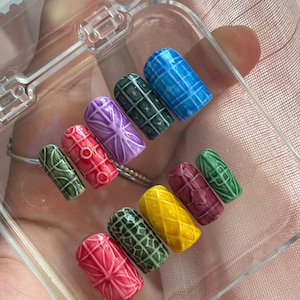 May include: A collection of colorful, textured artificial nails in a clear plastic case. The nails feature various designs, including geometric patterns and floral motifs, in shades of red, green, yellow, purple, and blue.