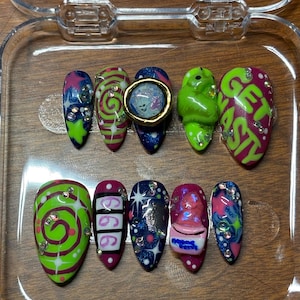 May include: A set of ten artificial nails with various designs. The nails feature vibrant colors like green, pink, blue, and purple, with embellishments such as rhinestones and 3D elements. Some nails have text that says "GET NASTY" and "666".