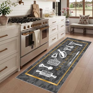 May include: A rectangular kitchen rug with a dark gray background and a gold border. White line drawings of kitchen utensils and the words "KITCHENWARE COOK AT STYLES" are featured on the rug. The rug is placed in front of a stainless steel oven.
