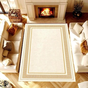 May include: A cream-coloured rectangular rug with a gold Greek key border. The rug is in a living room with white sofas, a fireplace, and a basket. The rug's dimensions are not visible.