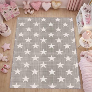 Op de afbeelding: Een rechthoekig grijs tapijt met een wit sterrenpatroon. Het tapijt ligt op een houten vloer in een kinderkamer. De kamer is ingericht met roze en witte accessoires, waaronder knuffels en een poppenhuis.