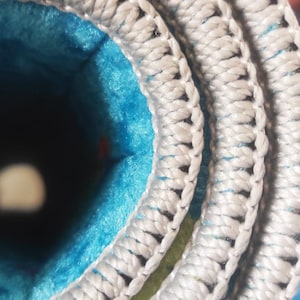 May include: A tightly rolled textile item featuring a bright blue and lime green patterned interior surface. The edges are finished with a thick, light gray braided or stitched border that creates a distinct layered spiral effect when viewed from the side.
