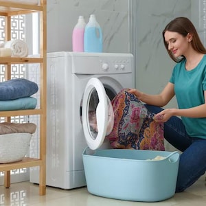 May include: A white front-loading washing machine with a circular door, a light blue plastic laundry basket, and a wooden shelving unit holding folded blue, light blue, and beige towels. Two detergent bottles sit atop the washing machine.
