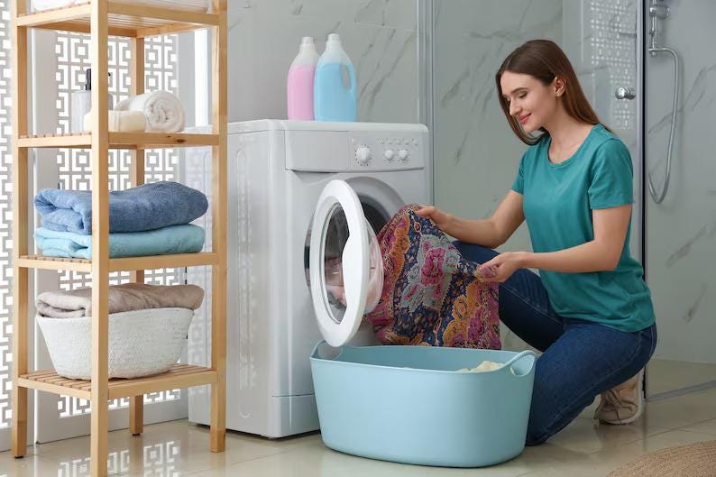 May include: A white front-loading washing machine with a circular door, a light blue plastic laundry basket, and a wooden shelving unit holding folded blue, light blue, and beige towels. Two detergent bottles sit atop the washing machine.