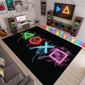 Op de afbeelding: Zwart rechthoekig vloerkleed met neon PlayStation-controller symbolen in groen, rood, blauw en paars. De symbolen zijn een driehoek, cirkel, kruis en vierkant, met een rookeffect. Het vloerkleed ligt in een gamekamer.