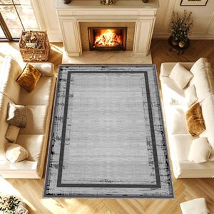 Puede incluir: Una alfombra rectangular con un centro gris claro y un borde gris oscuro. La alfombra tiene un aspecto desgastado y texturizado, y está colocada en un salón con sofás blancos, una chimenea y una cesta.