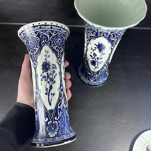 May include: Two white and blue ceramic vases with floral designs. The vases have a flared base and a wide opening. The blue patterns include flowers and ornate scrollwork. One vase is held by a person.